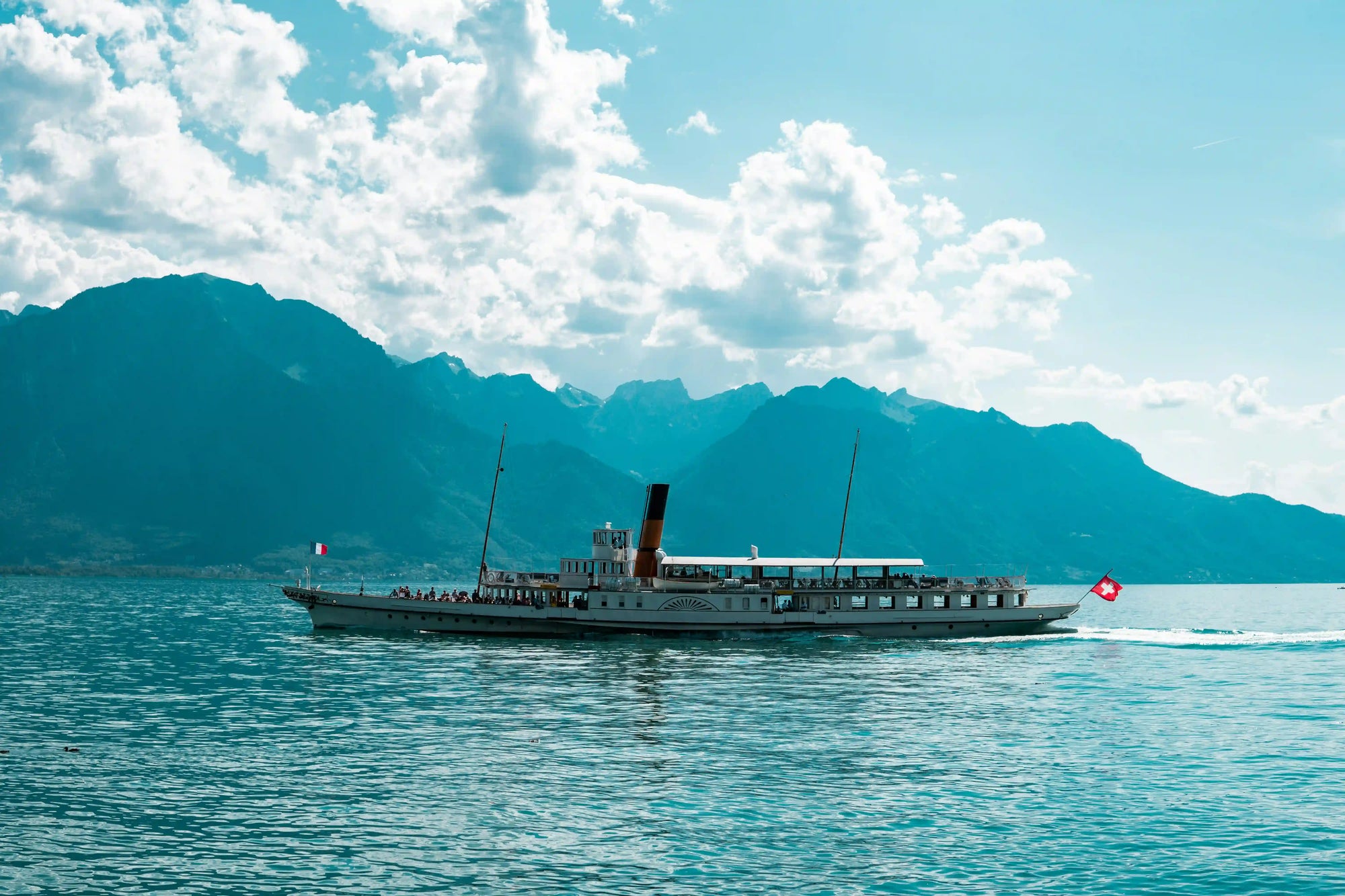 Bateau à roue naviguant sur le lac Léman avec les drapeaux français et suisse, illustrant les origines franco-suisses de la Maison AYNAUD.